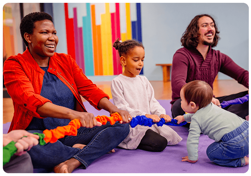Families with arms outstretched touching a stretchy band with baby moving up to other child and touching band together